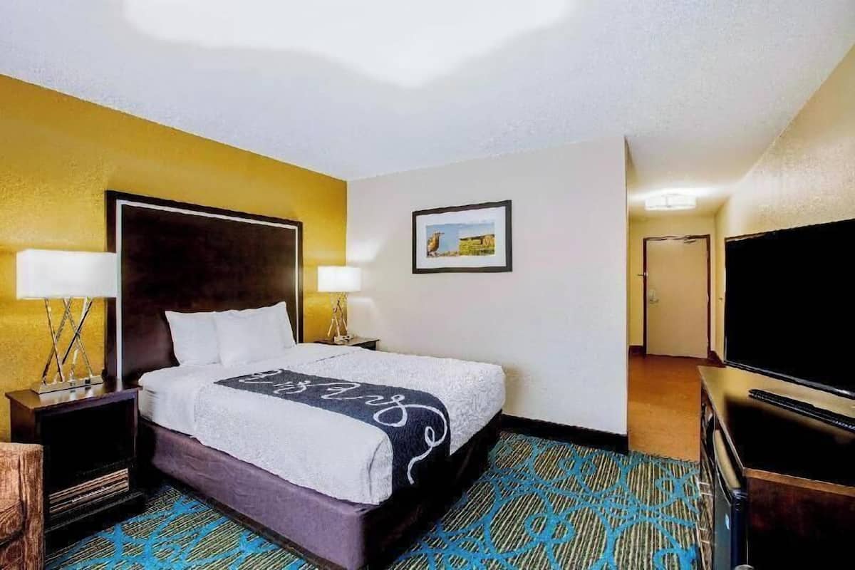 Ein Hotelzimmer mit einem Bett, Nachttischlampen, einem Flachbildfernseher, einem gerahmten Bild an der Wand und einem Teppichboden.