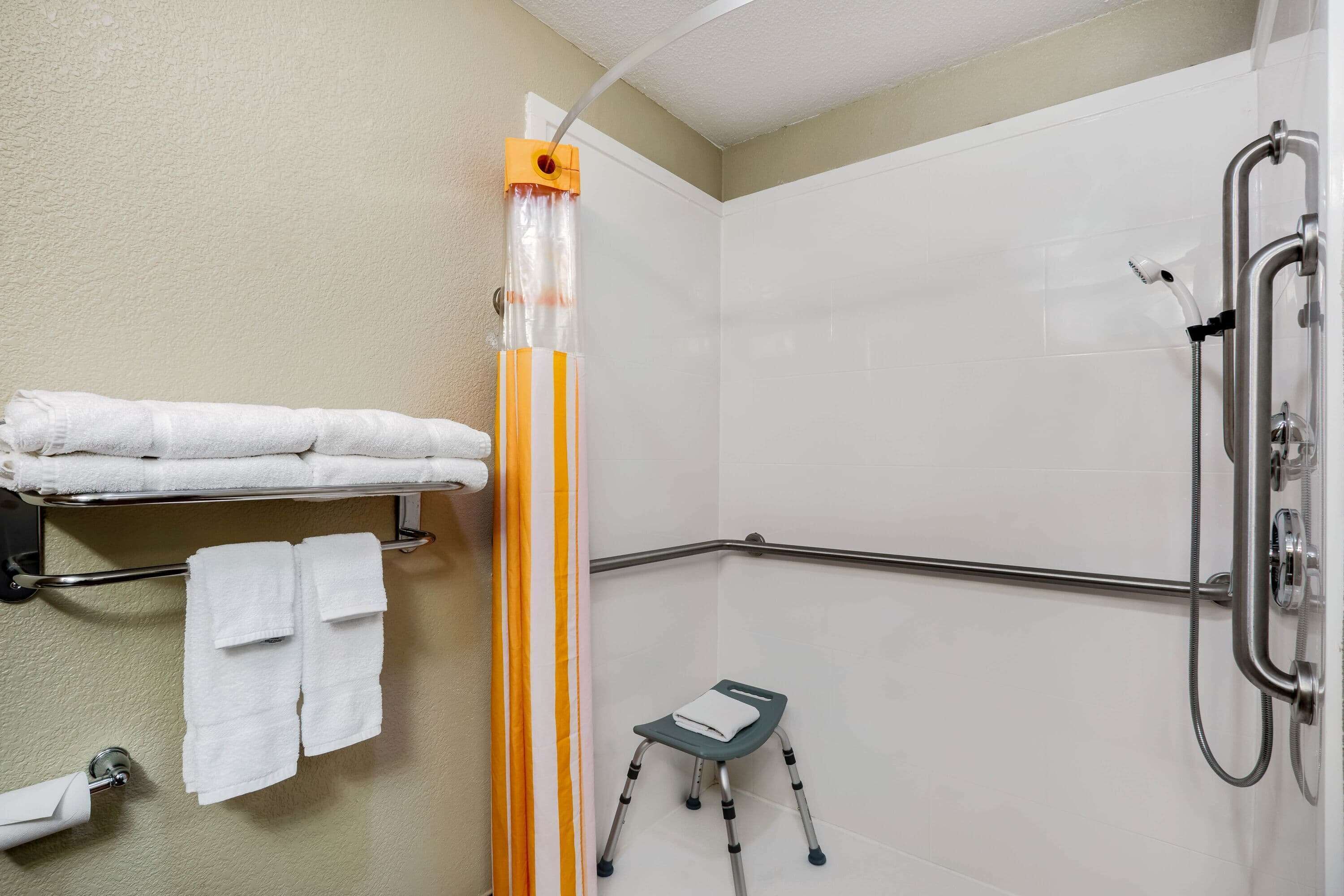 Un baño con ducha, un perchero con toallas, un taburete y una manguera de ducha.