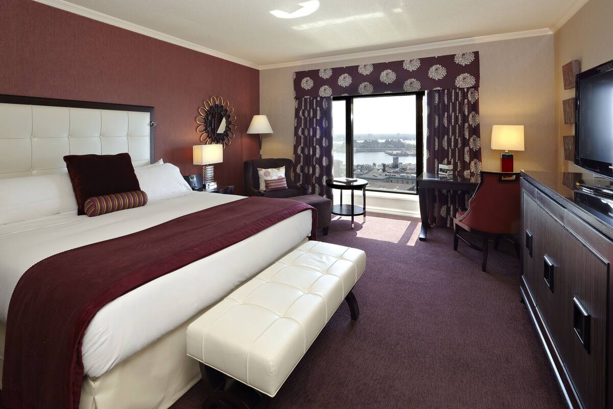 A hotel room with a large bed, a desk, a chair, a TV, and a view of the city.