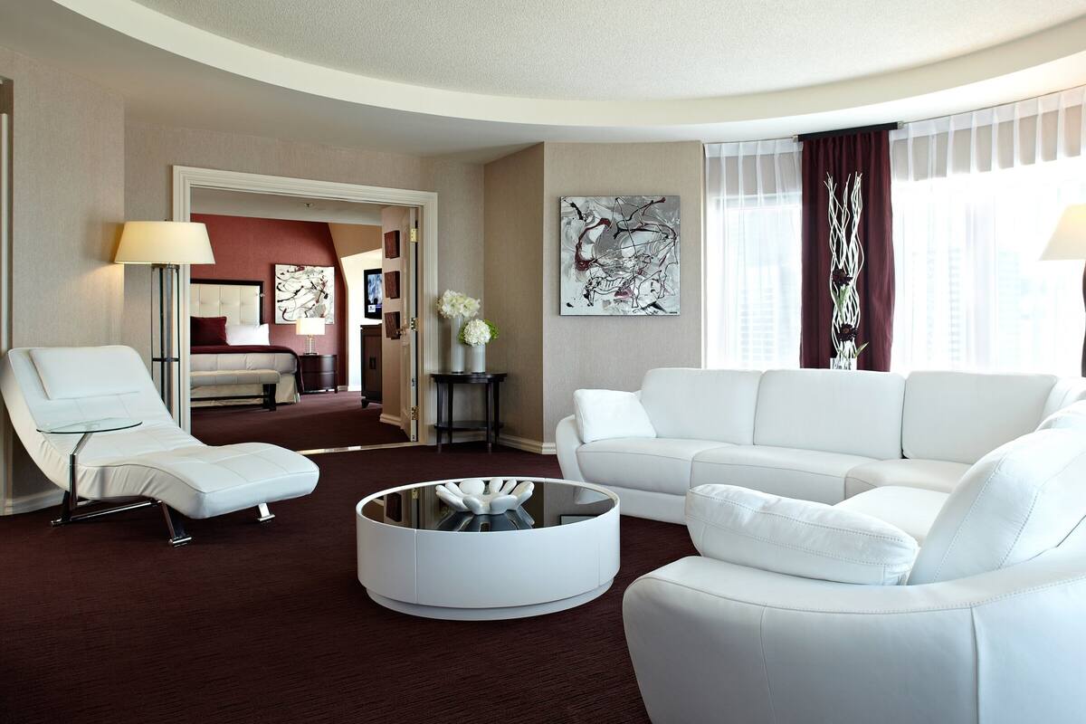 A modern hotel room with a white sofa set, a lounge chair, a round coffee table, and a view of the city through large windows.