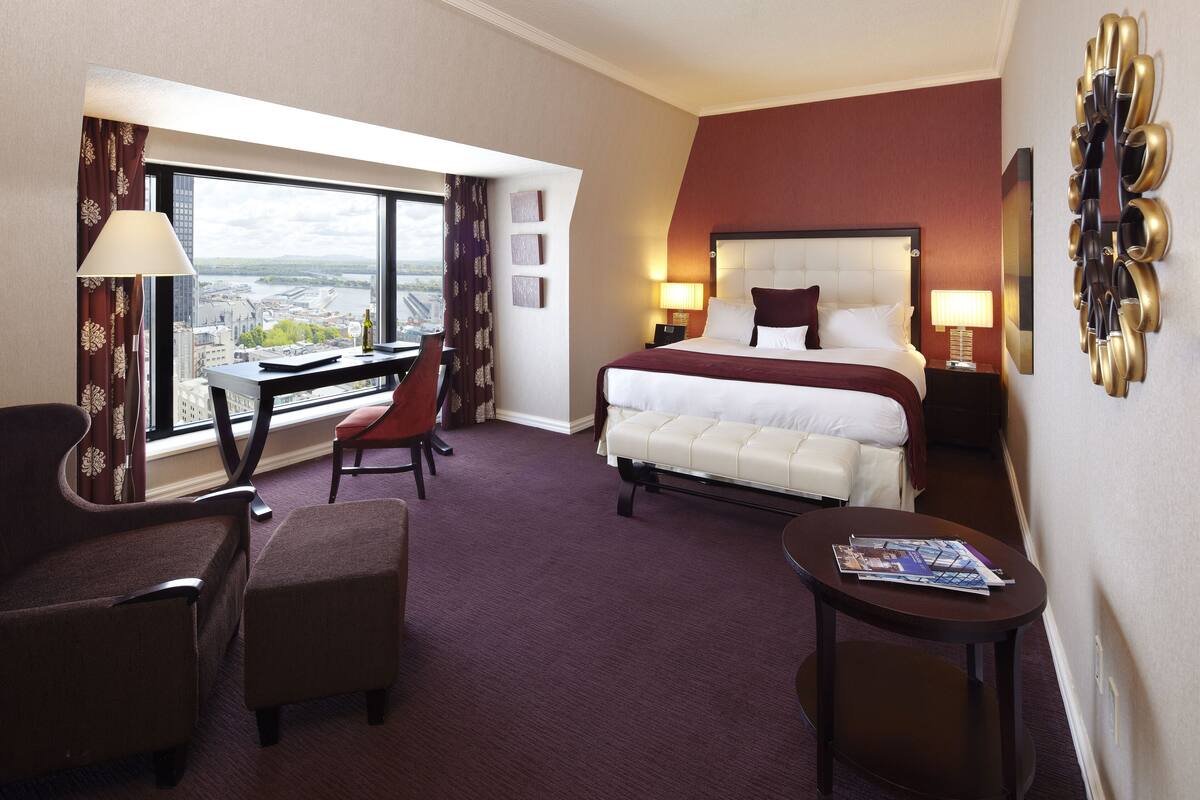 A hotel room with a large bed, a desk, a chair, and a view of a cityscape.