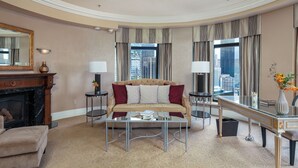 A hotel room with a sofa, two side tables, a coffee table, a fireplace, a mirror, and a view of the city.