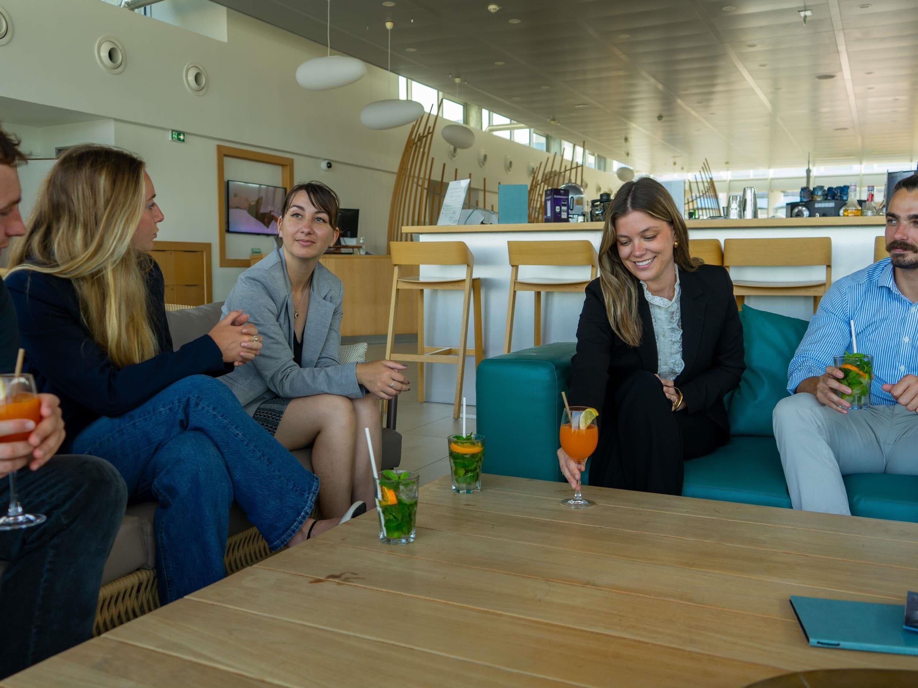 A group of people seated at a wooden table in a modern interior with a bar area in the background.