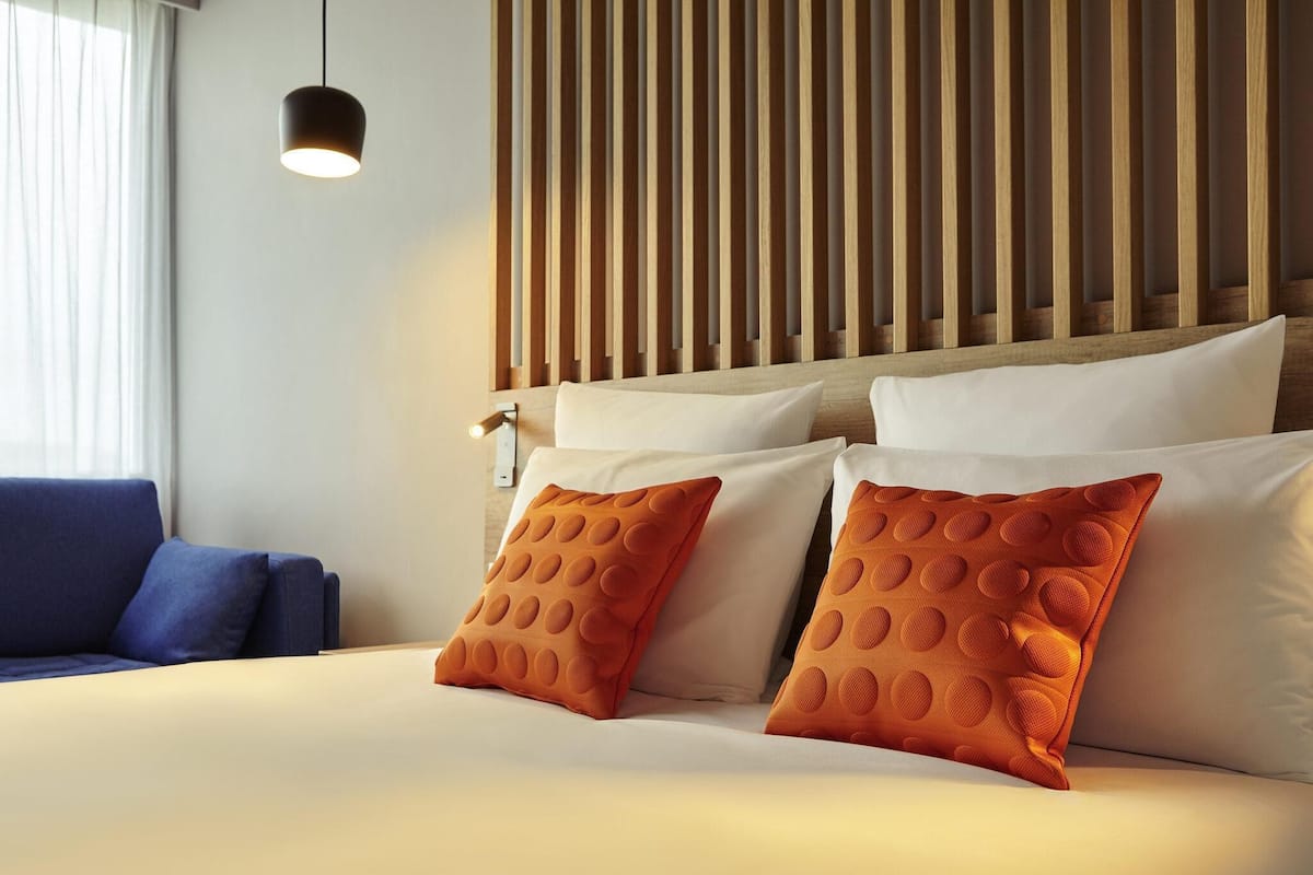 A hotel room with a bed, two pillows, a blue sofa, a wooden slat headboard, and a pendant light.