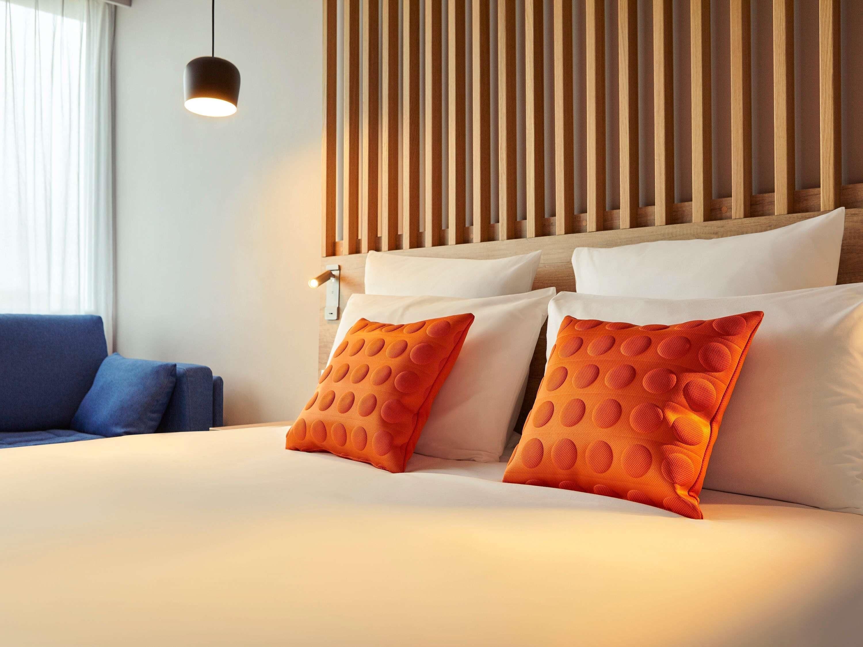 Habitación de hotel con cama, dos almohadas, un sofá azul, cabecera de madera con listones y una lámpara colgante.