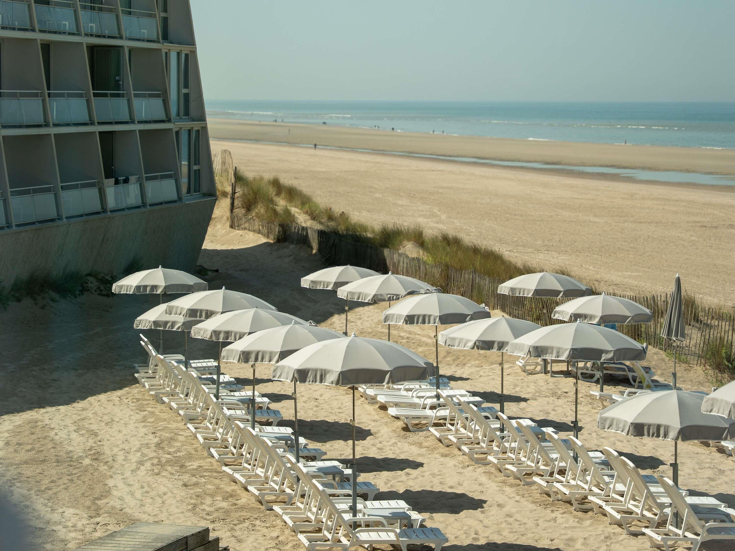 A beachside hotel with rows of sun loungers and umbrellas, a sandy beach, and the ocean in the background.
