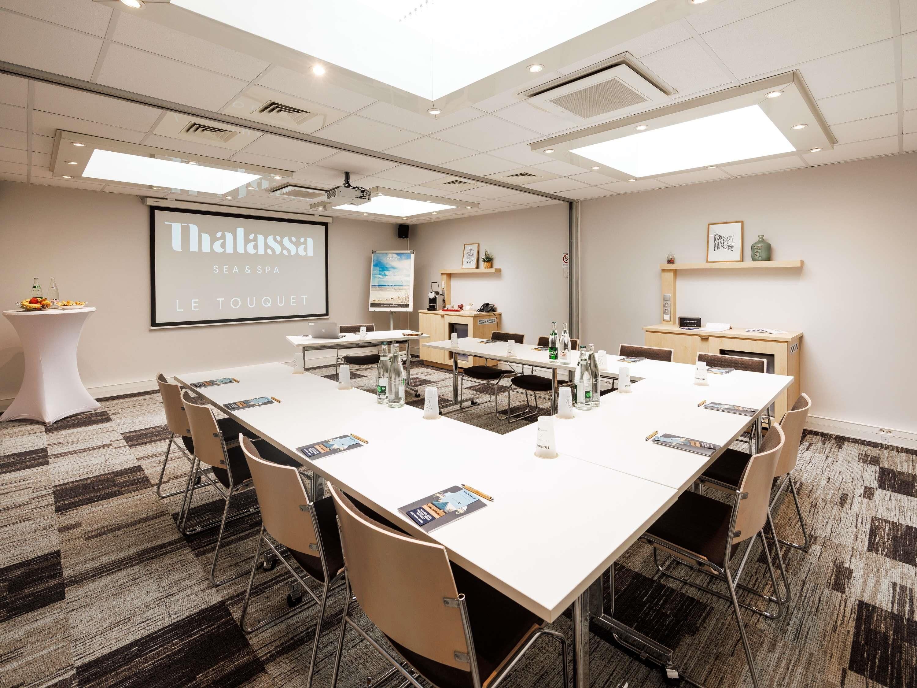 Un salón de conferencias con una larga mesa, sillas, una pantalla de proyector y una presentación sobre Thalassa Sea & Spa.