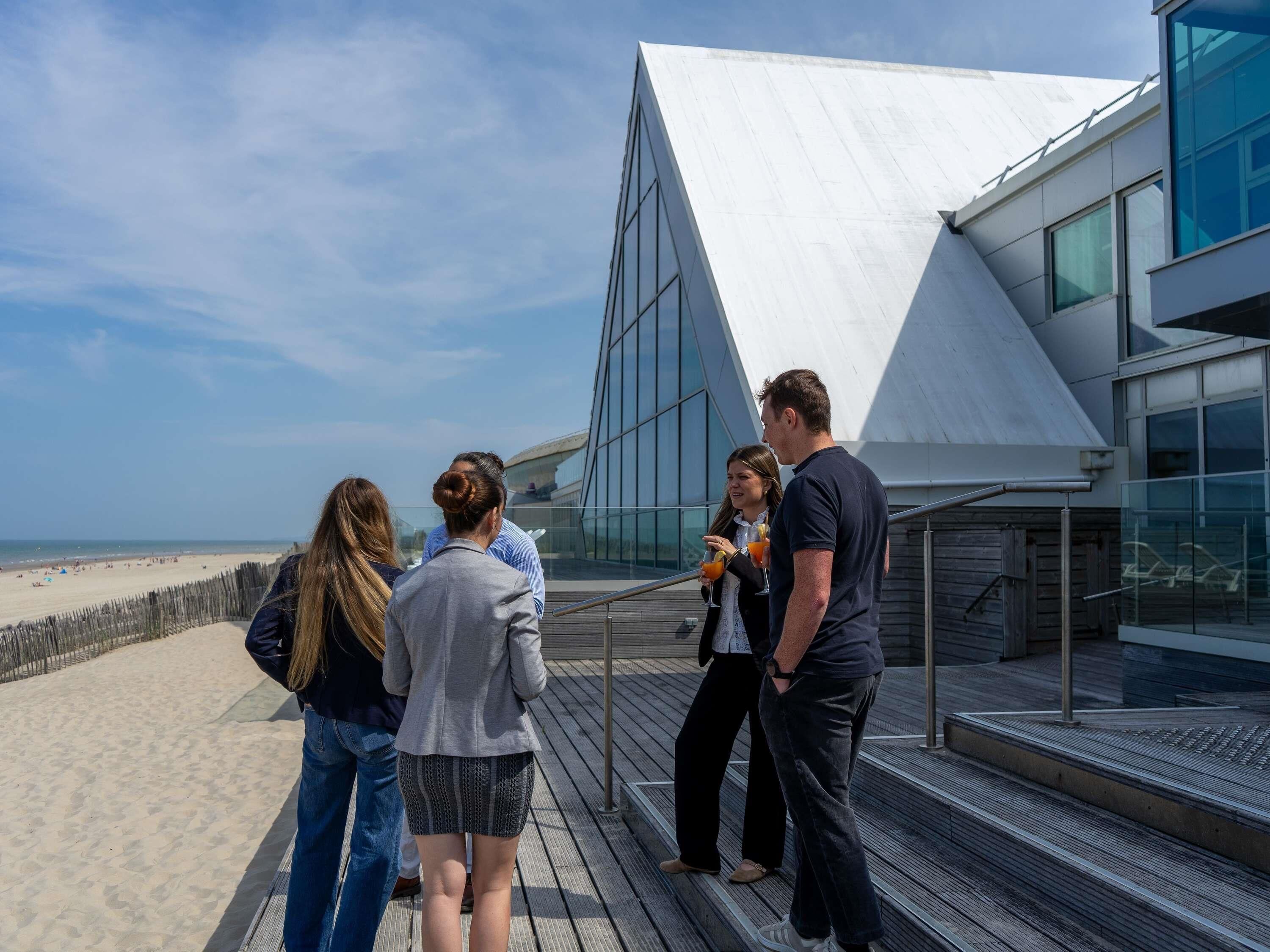 Eine Gruppe von Menschen steht auf einem Holzdeck mit Blick auf einen Strand, im Hintergrund ein modernes Gebäude mit großen Fenstern und einem geneigten Dach.