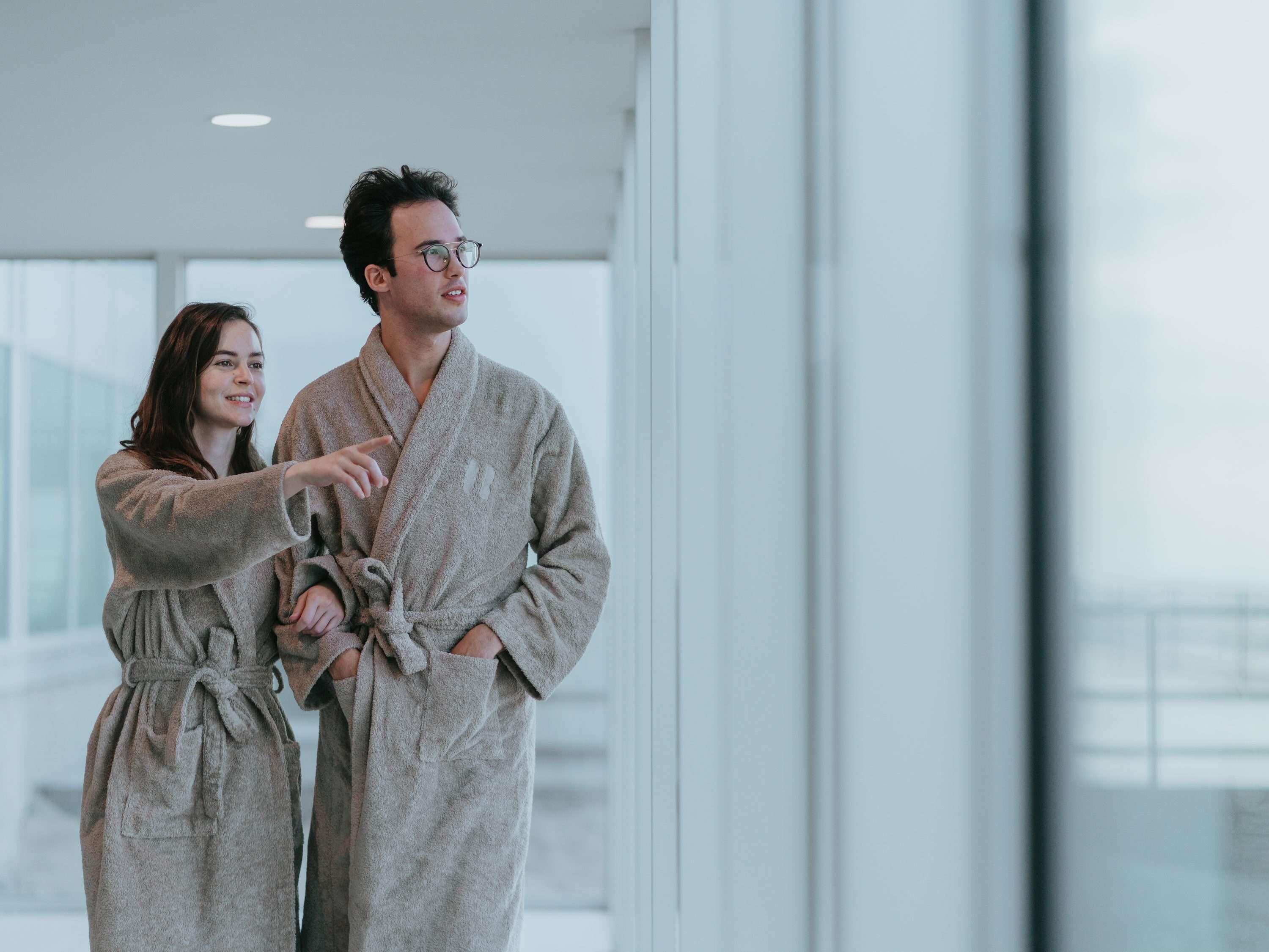 A man and a woman in matching bathrobes standing in a modern hotel lobby with large windows.