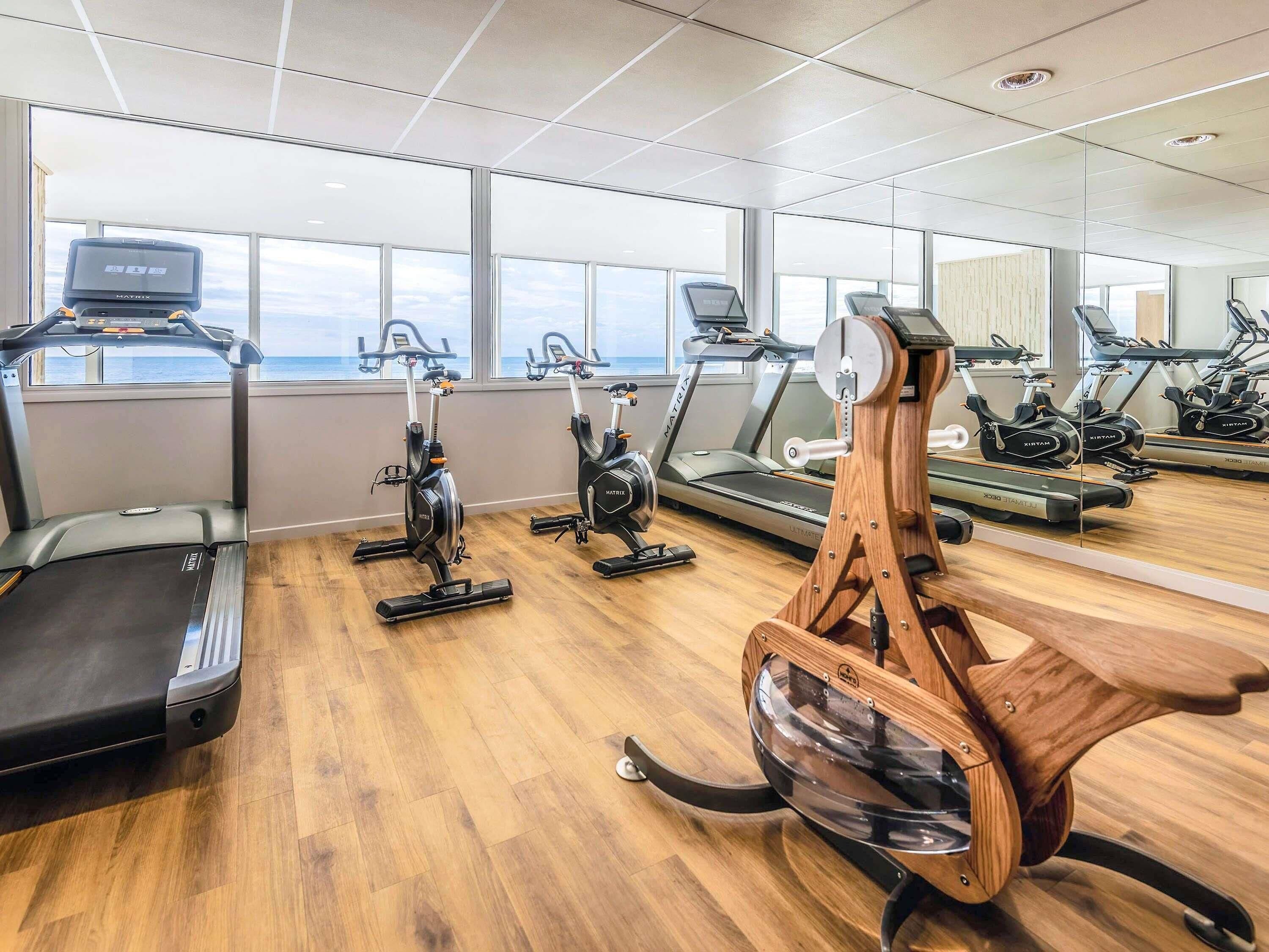Une salle de sport équipée de nombreux appareils d’exercice, notamment des tapis de course et des vélos d’appartement, et dotée de grandes fenêtres laissant entrer la lumière naturelle.