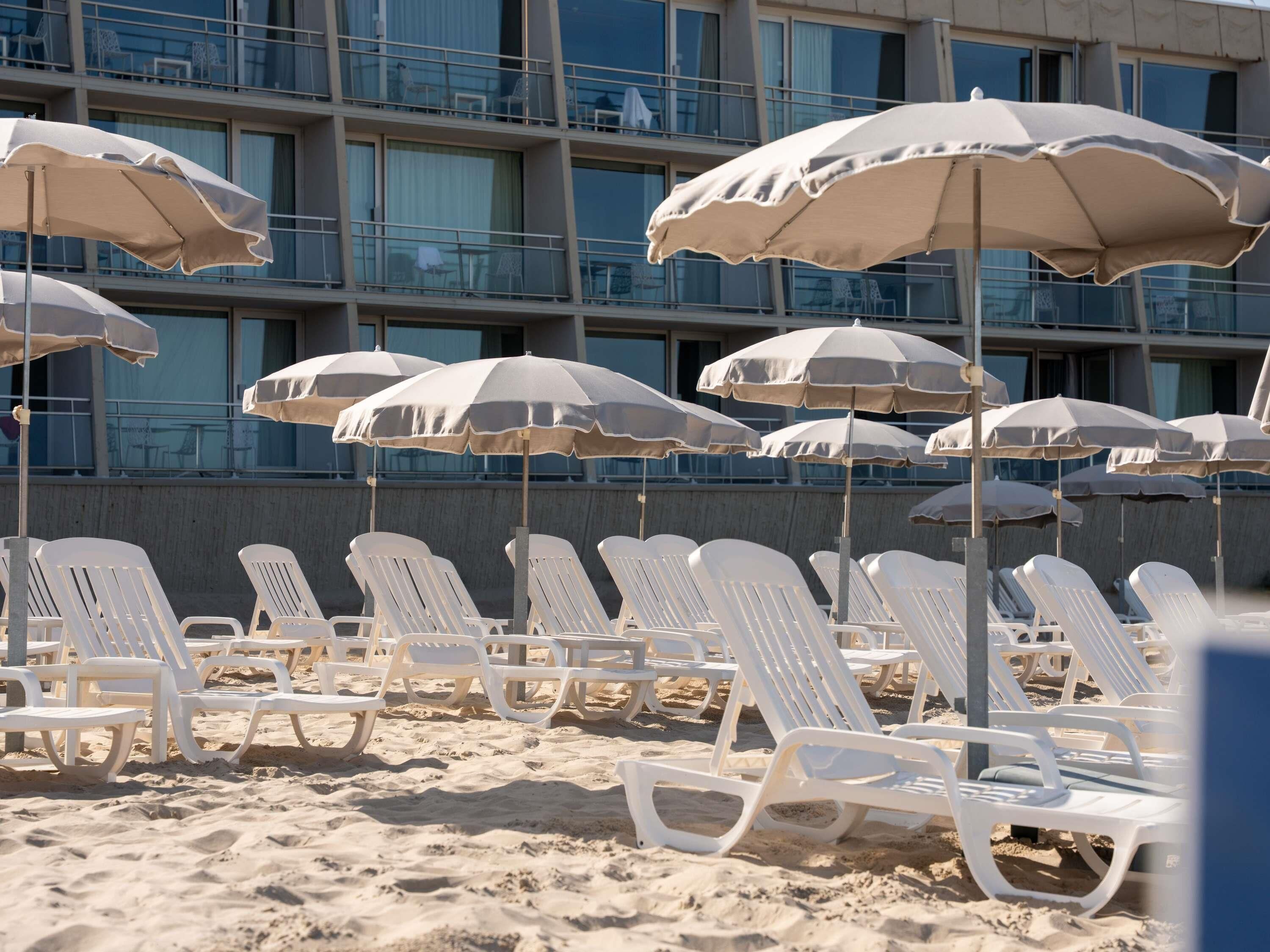 Una playa con sillas y sombrillas blancas, y un edificio con balcones al fondo.