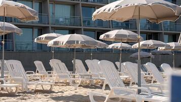 Una playa con sillas y sombrillas blancas, y un edificio con balcones al fondo.