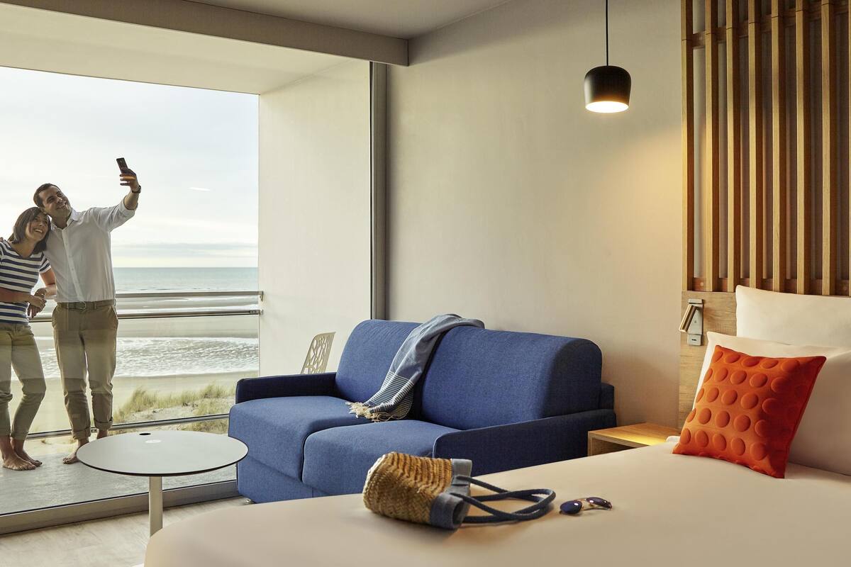Ein modernes Hotelzimmer mit einem großen Fenster, einem blauen Sofa, einem Bett mit einem orangefarbenen Kissen und einer hängenden Pendelleuchte.