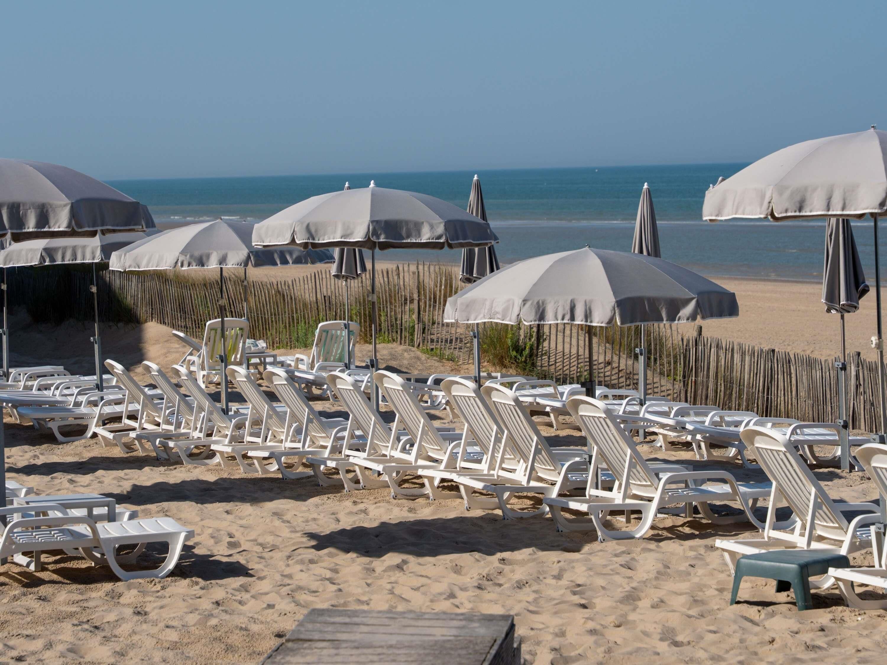 A beach with rows of white lounge chairs and grey umbrellas, a wooden fence, and the ocean in the background.