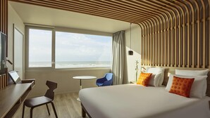 A hotel room with a large bed, a desk, a chair, a small table, and a view of the ocean. - Novotel Thalassa Le Touquet Hotel (Le Touquet-Paris-Plage)
