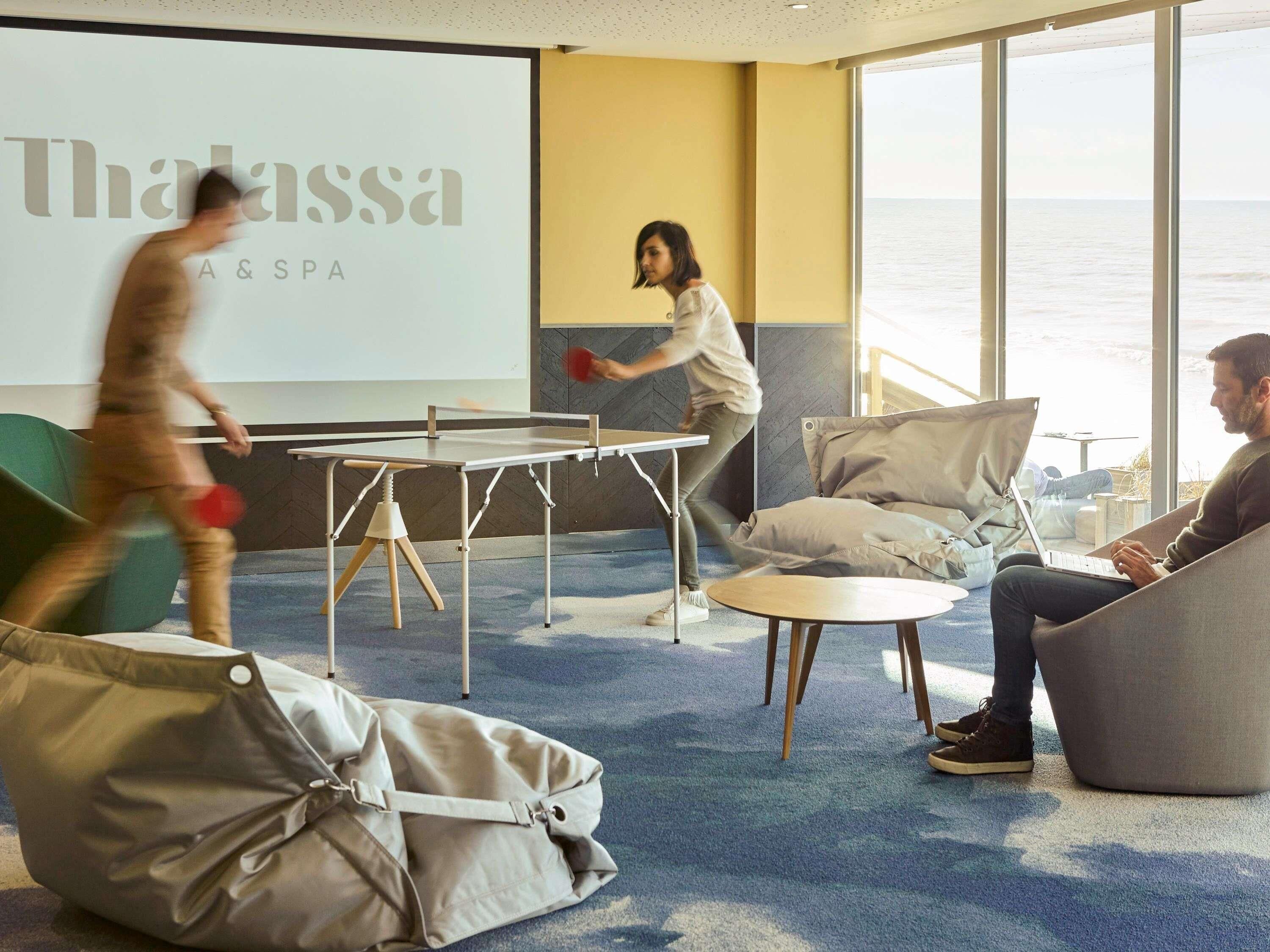 A modern lounge area with a ping pong table, comfortable seating, and a large screen displaying the name 'Thalassa SPA'.