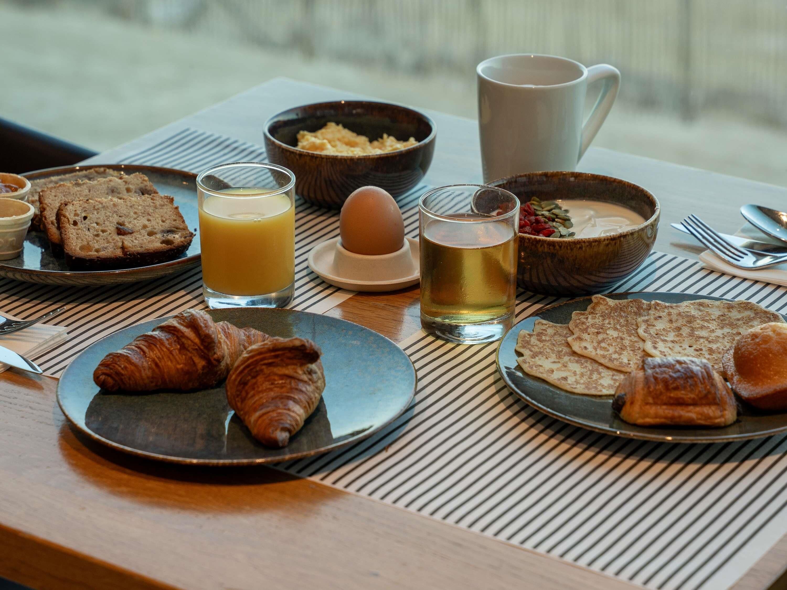 Una mesa de desayuno con cruasanes, tostadas, un vaso de zumo de naranja, una taza de té y un cuenco de cereales.