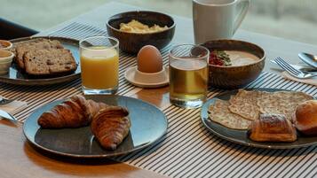 Una mesa de desayuno con cruasanes, tostadas, un vaso de zumo de naranja, una taza de té y un cuenco de cereales.