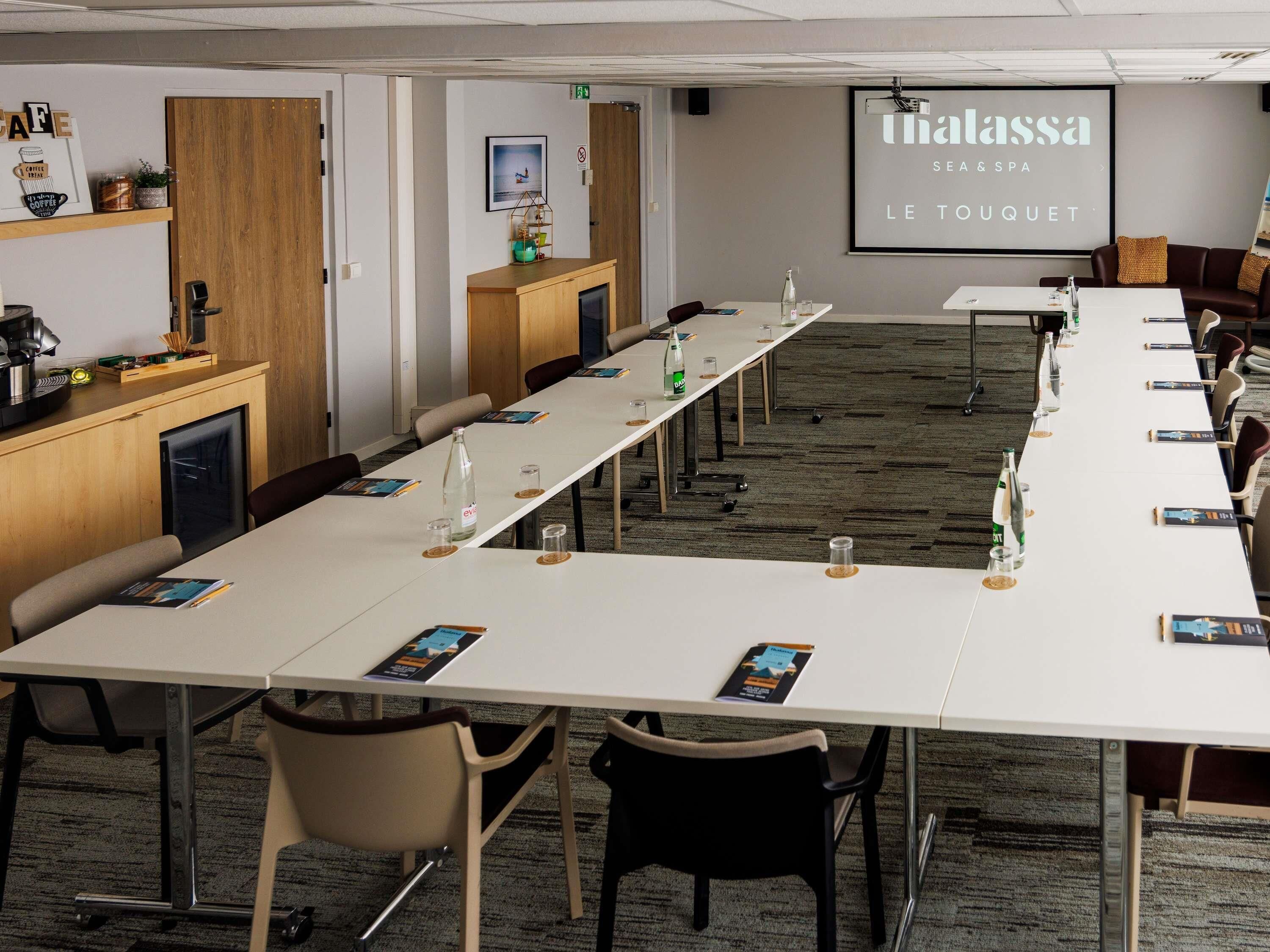 A conference room with a long table, chairs, a coffee station, and a screen displaying 'Thalassa SEA & SPA LE TOUQUET'.