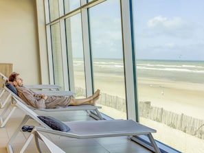 A person lying on a sun lounger with a view of the beach and ocean through large windows.