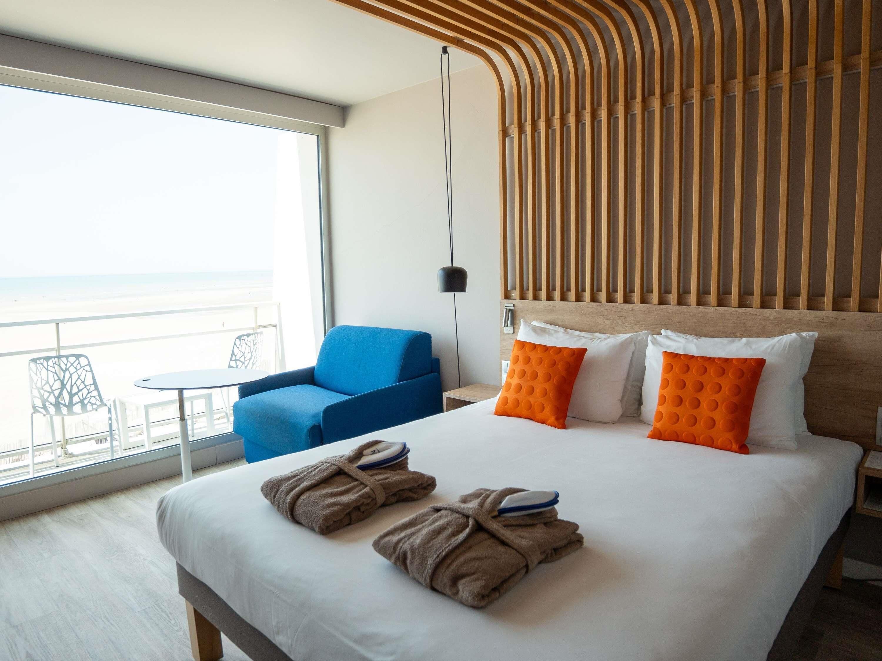 Habitación de hotel con cama, una silla azul, balcón y vistas, y cabecera de madera.