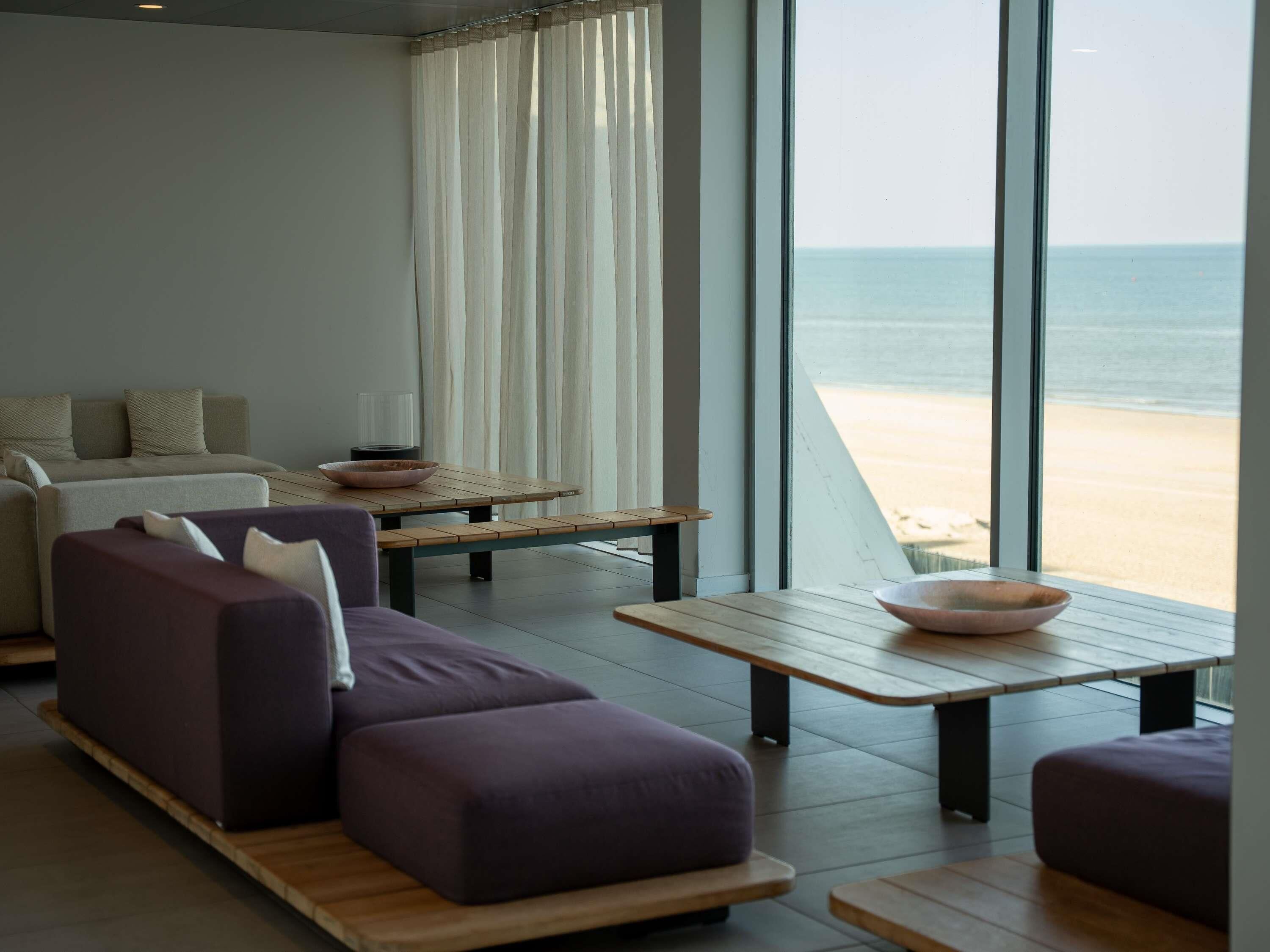 A modern living room with a view of the beach, featuring a sofa, a table with a bowl, and a large window.