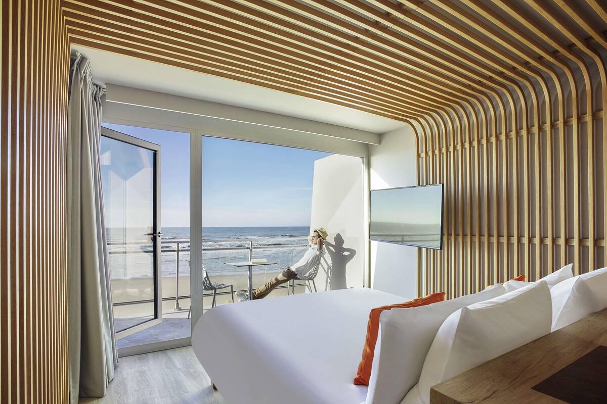 Ein modernes Hotelzimmer mit einem großen Bett, einem Fernseher und einem Balkon mit Blick auf den Strand.