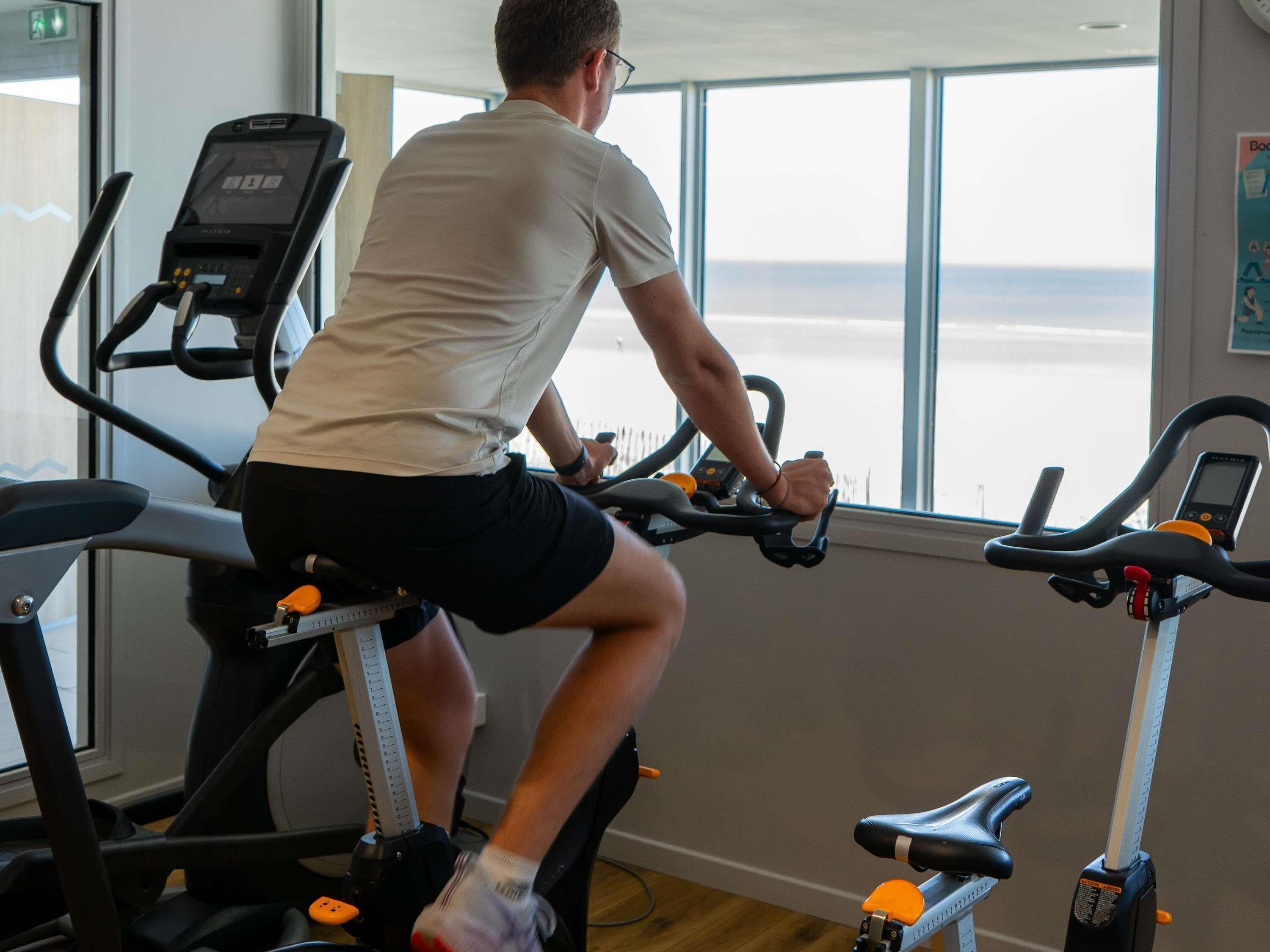 A person using an exercise bike in a room with large windows and a treadmill.