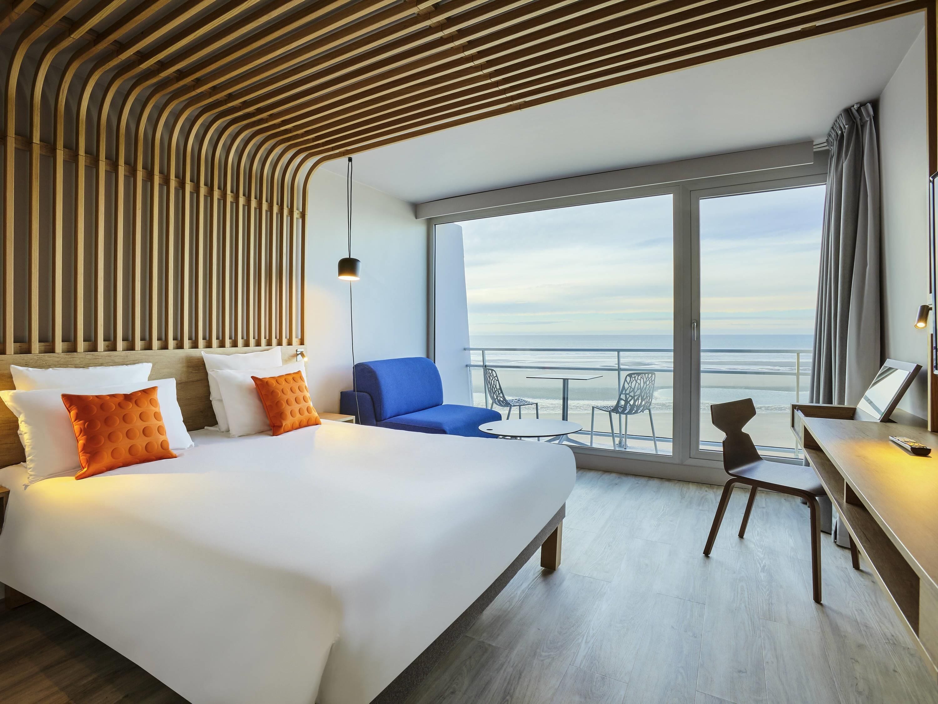 A hotel room with a bed, a desk, a chair, and a balcony with a view of the ocean.