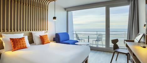 Quarto de hotel com cama, escrivaninha, cadeira e varanda com vista para o oceano.