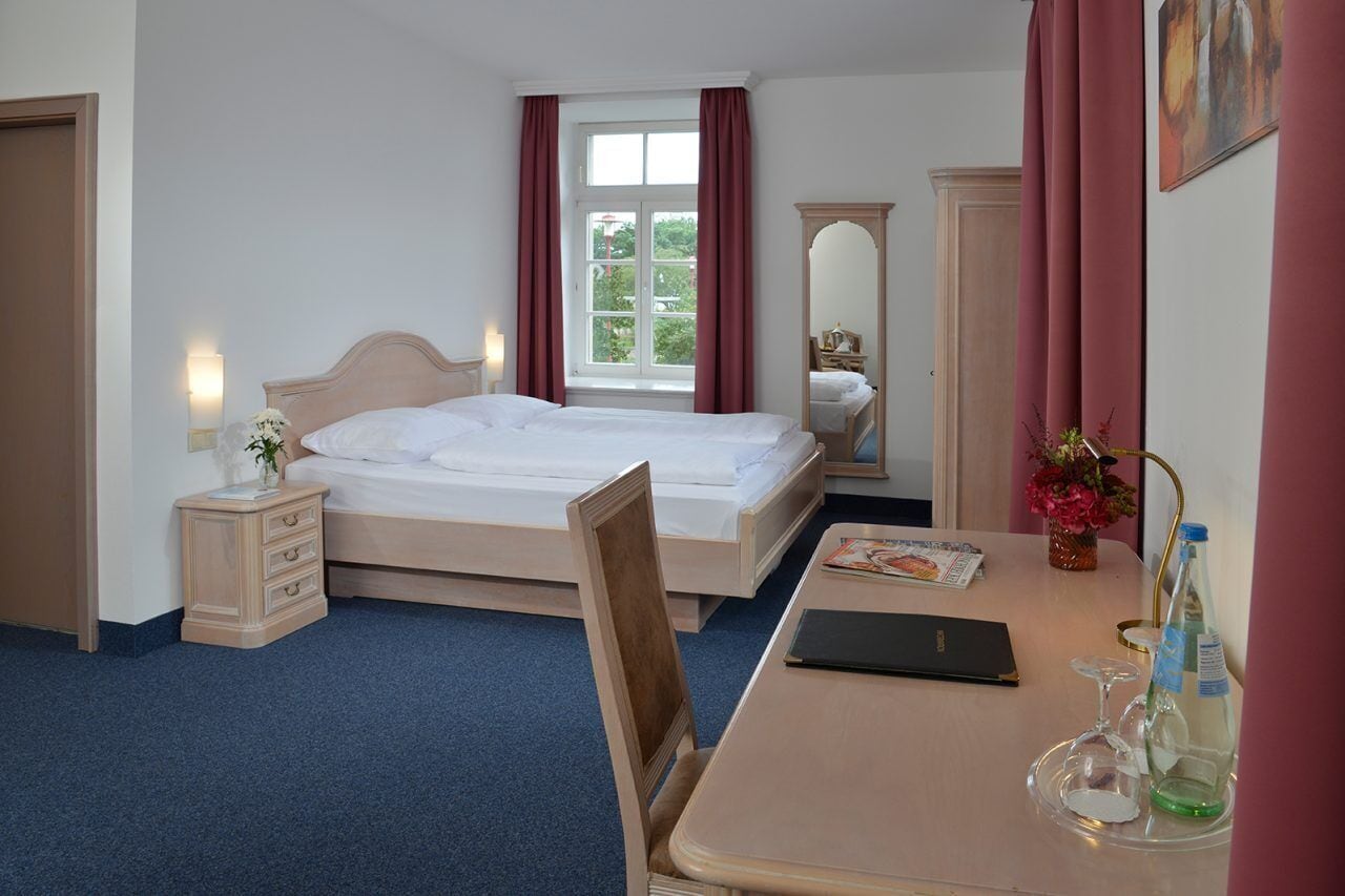 Une chambre d’hôtel avec un lit simple, une table de chevet avec une lampe, une chaise, une petite table avec un magazine et une fenêtre avec des rideaux.