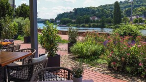 An outdoor seating area with wicker furniture and potted plants, overlooking a river and a bridge. - Hotel Stadt Hameln (Hamelin)