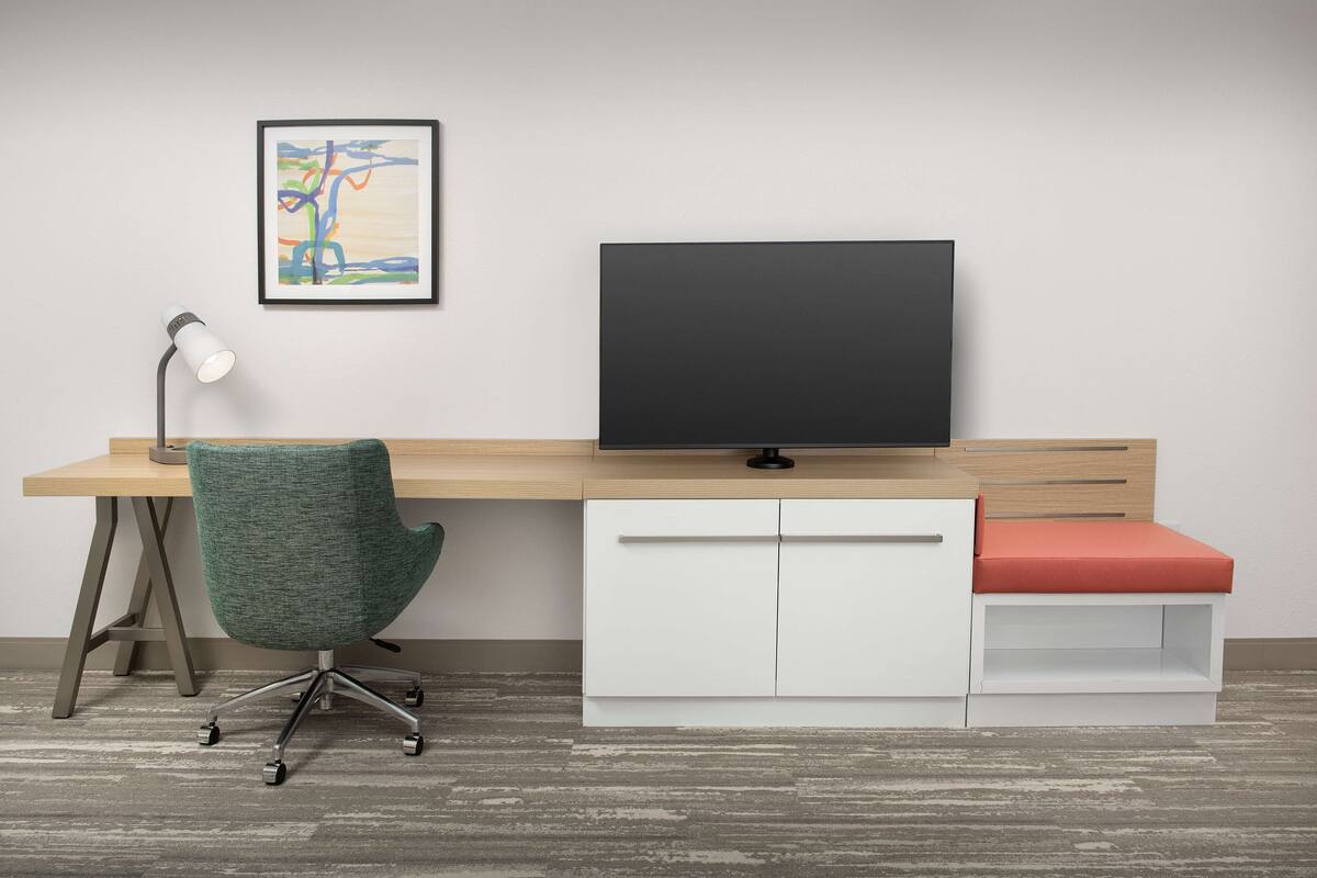 A modern office desk with a flat-screen monitor, a green office chair, a white storage unit, and a red cushioned bench.