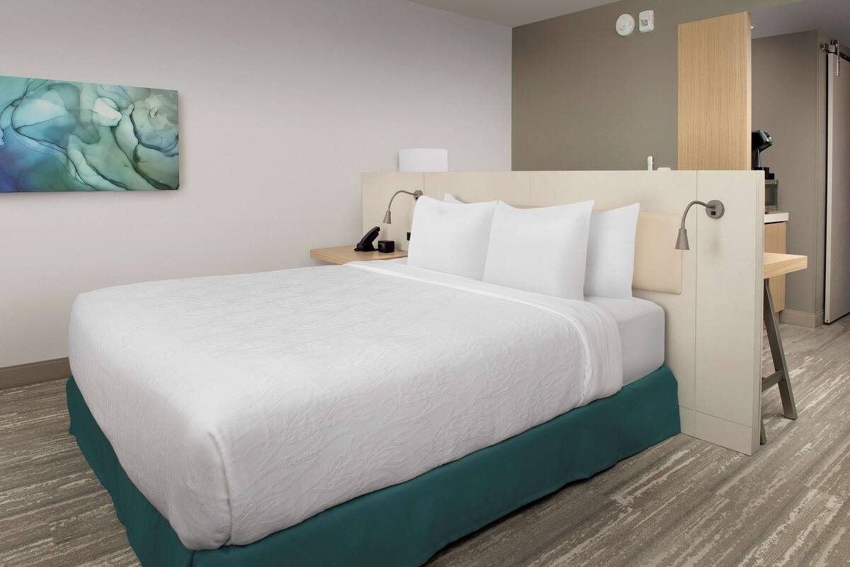 A neatly made bed with white linens, a teal bed skirt, and a wooden headboard. There is a bedside table with a lamp, a small nightstand, and a wall-mounted shelf.