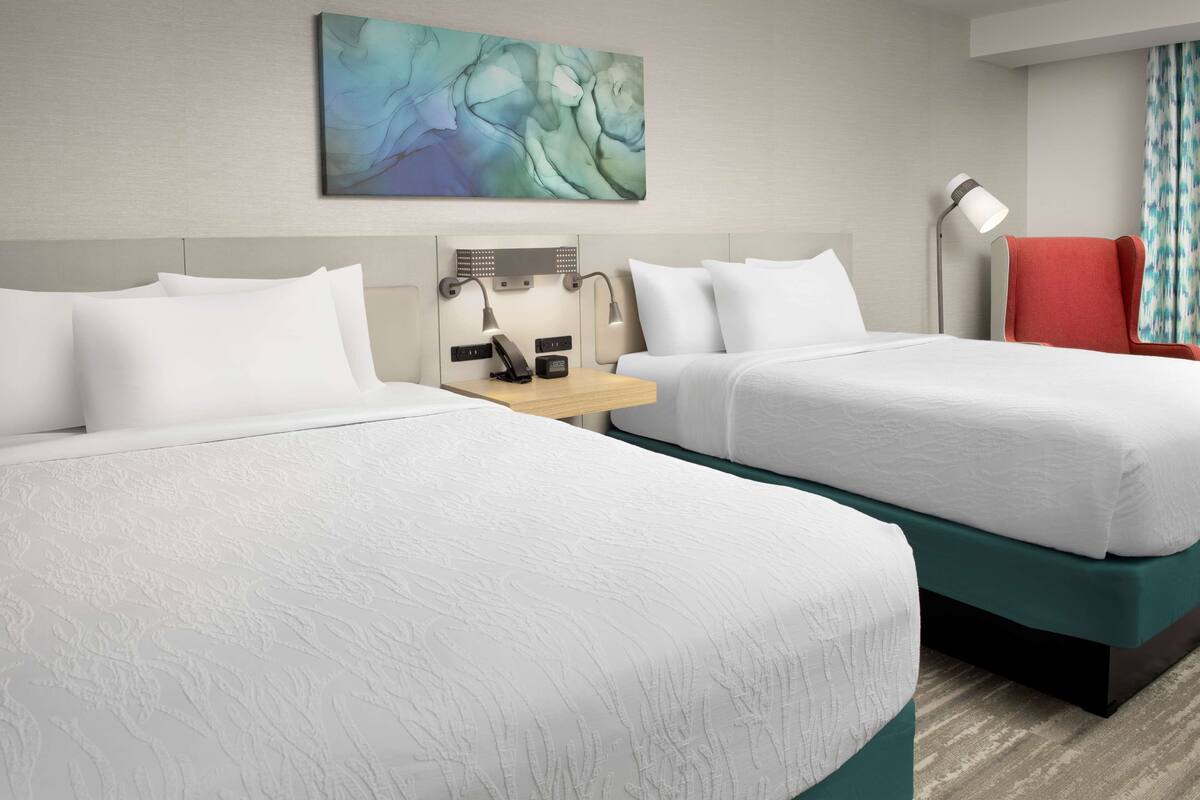 A hotel room with two beds, a desk with a lamp, a red chair, and a wall-mounted artwork.