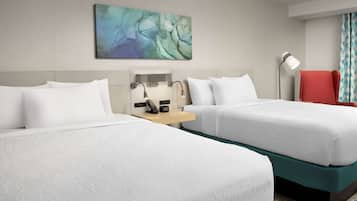 A hotel room with two beds, a desk with a lamp, a red chair, and a wall-mounted artwork.
