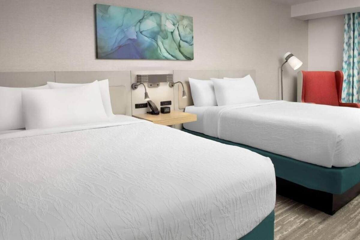 A hotel room with two beds, a desk with a lamp, a red chair, and a painting on the wall.