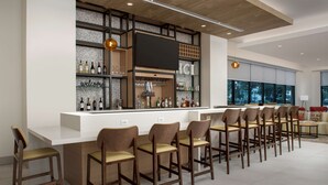 A modern bar area with a long counter, multiple bar stools, and a variety of bottles and glasses. - Hilton Garden Inn Orlando Downtown (Orlando)