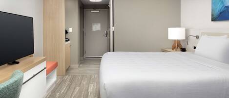 A hotel room with a bed, a TV, a desk, a chair, a nightstand, and a painting on the wall.