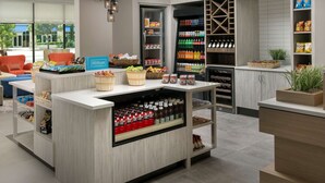 A modern hotel lobby with a central refreshment station stocked with snacks, beverages, and a minibar. - Hilton Garden Inn Orlando Downtown (Orlando)