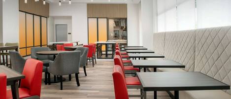 Un restaurante moderno con mesas altas y sillas rojas, un amplio banco acolchado y una cocina al fondo.