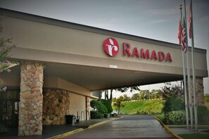 A Ramada hotel entrance with a stone pillar, a covered walkway, and a parking area.