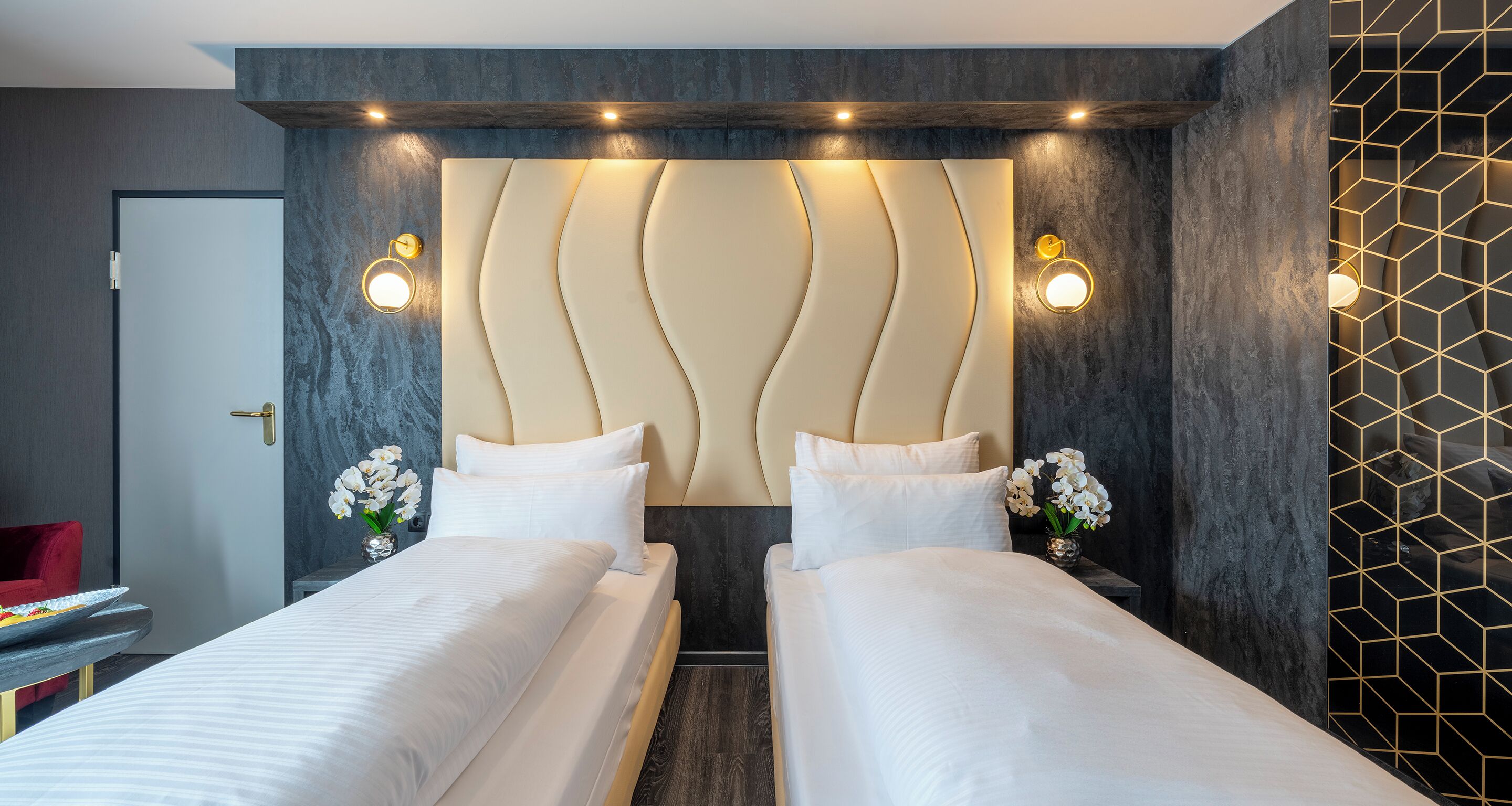 A hotel room with two beds, a modern headboard, wall sconces, and a geometric-patterned wall.