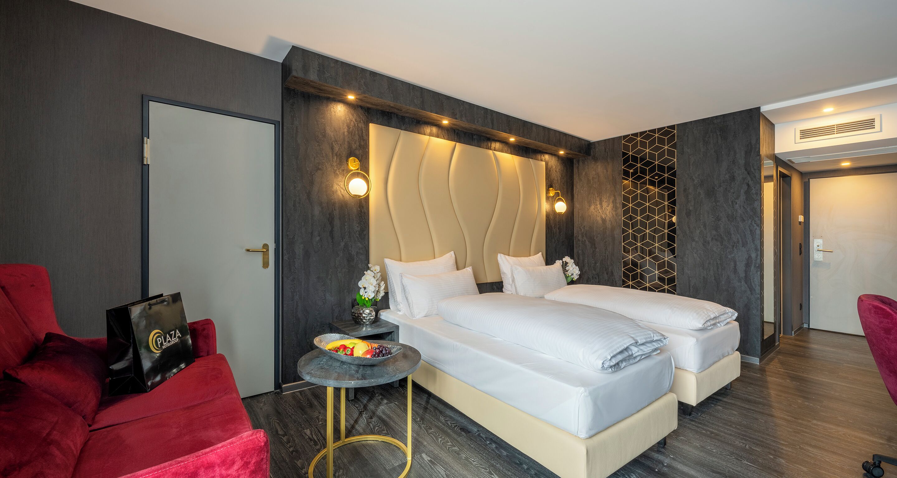 Une chambre d’hôtel moderne avec un lit double, une petite table ronde sur laquelle se trouve un saladier de fruits, un fauteuil rouge et un canapé rouge.