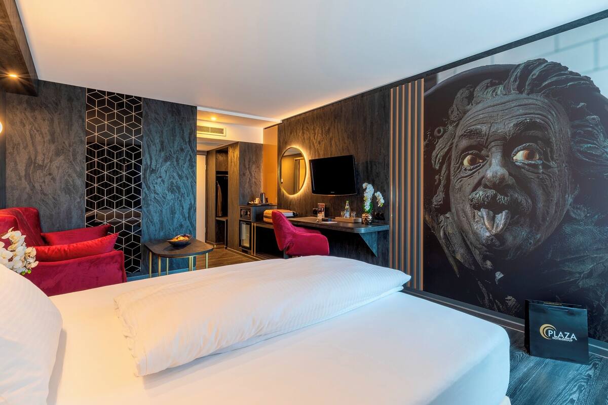 Ein Hotelzimmer mit einem großen Bett, roten Sesseln, einem Schreibtisch, einem Fernseher und einem Wandbild.