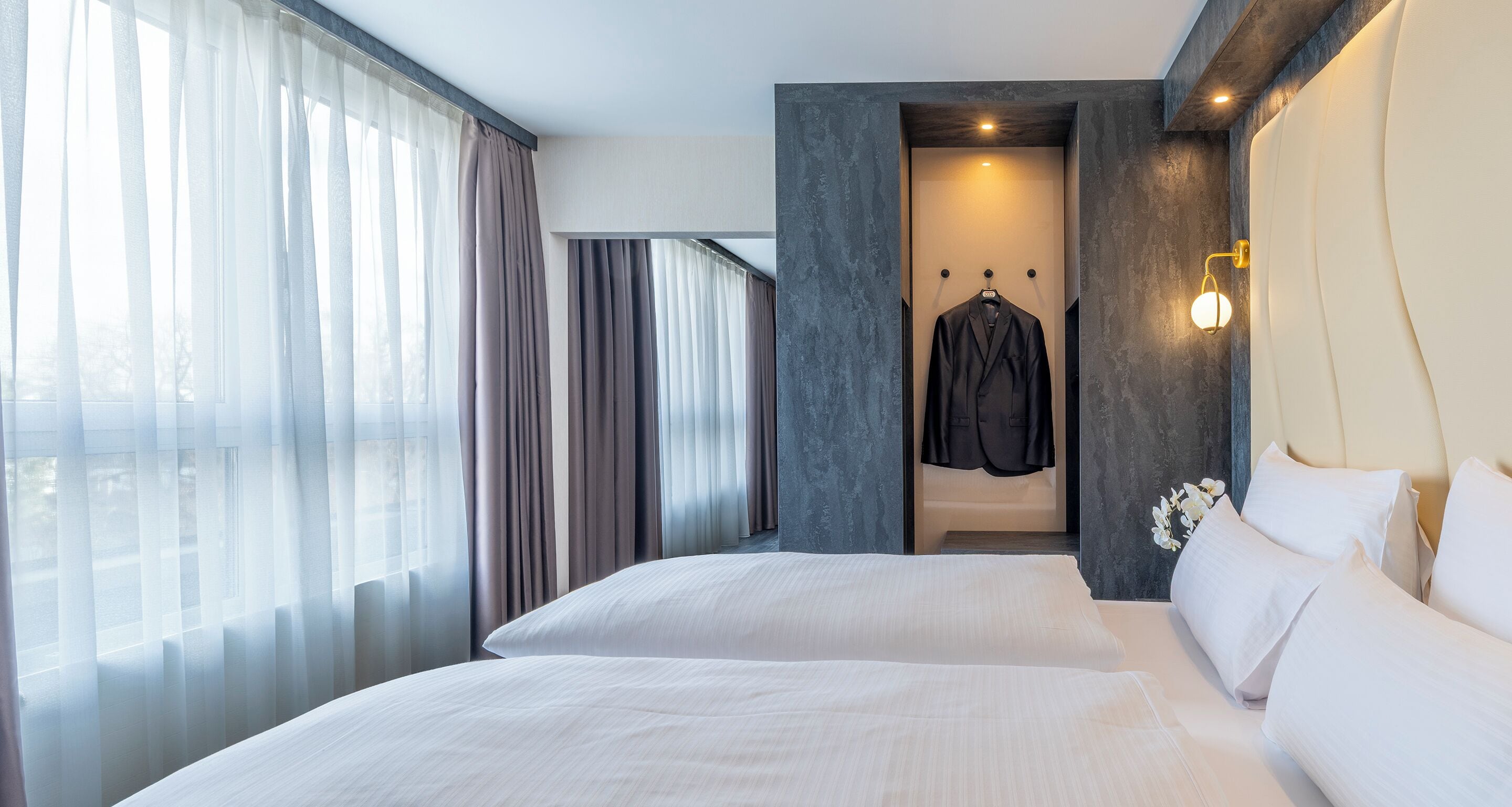 A modern hotel room with a large bed, a walk-in closet, and a view of the city through sheer curtains.