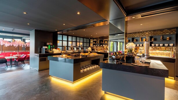 A modern restaurant with a central island bar, seating area with tables and chairs, and a view of the city through large windows.