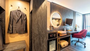 A modern hotel room with a grey suit hanging on the wall, a round mirror, a desk with a red chair, and a minibar.