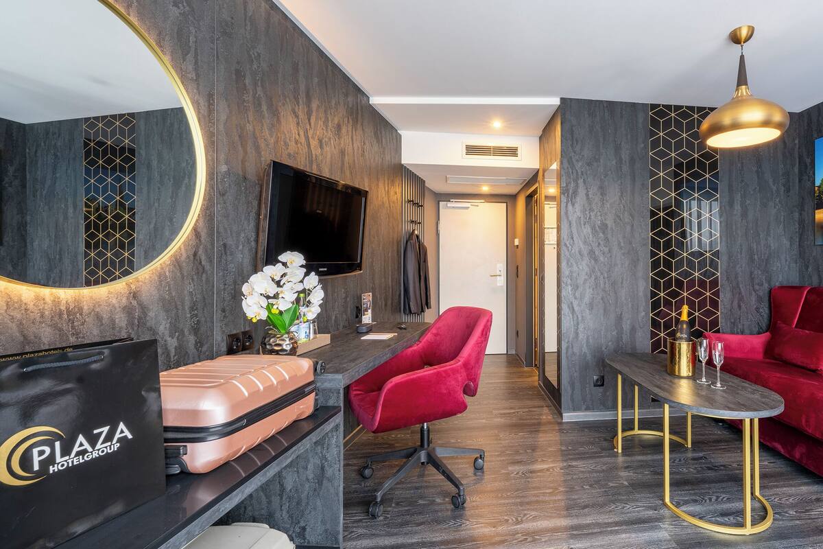 Ein modernes Hotelzimmer mit dunklem Holzboden, einem roten Samtsofa, einem Schreibtisch mit goldener Lampe und einem großen Stadtbild.