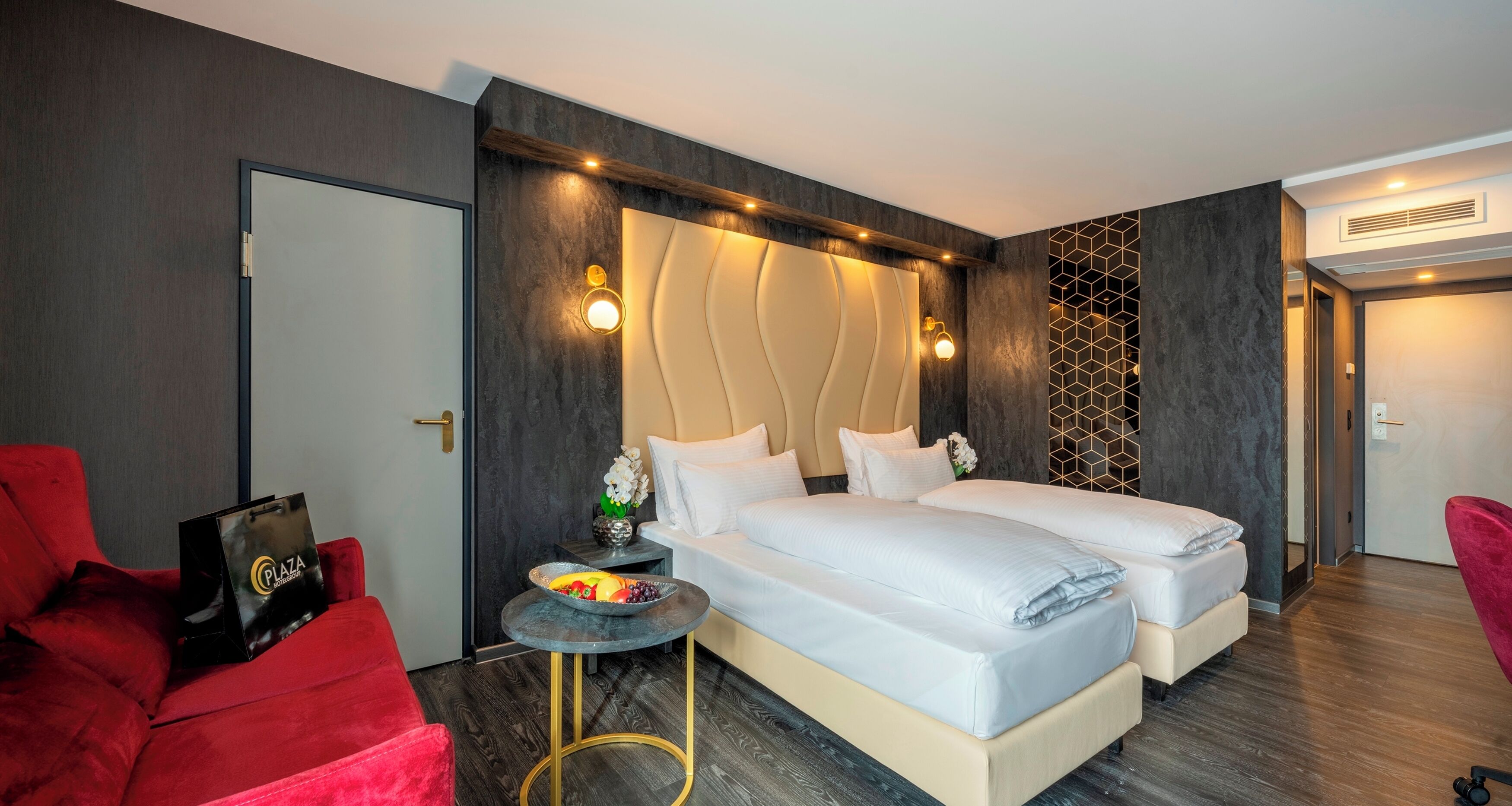 A modern hotel room with two beds, a red armchair, a small round table with a fruit bowl, and a decorative wall panel.